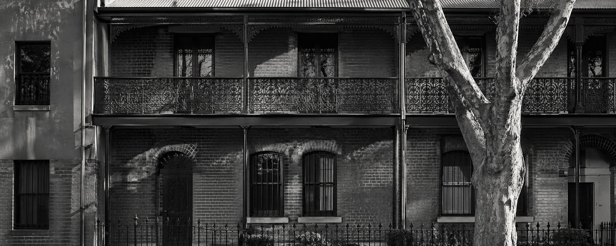 Inner-suburb terrace facade — brick, cast-iron lacework balcony, arched entries, mature plane tree casting dappled shadow across the rendered wall